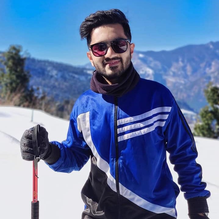 Shoumik, a young man, similing at the camrera. He is wearing a blue-black tracksuit, a sunglass, holding a red pole. Background shows a snowy mountain of Uttarakhand, India.